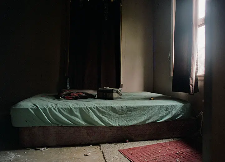 An abandoned room at Lily of the Valley Campground with a bed, light green sheet, dark curtains, and a sunlit window.