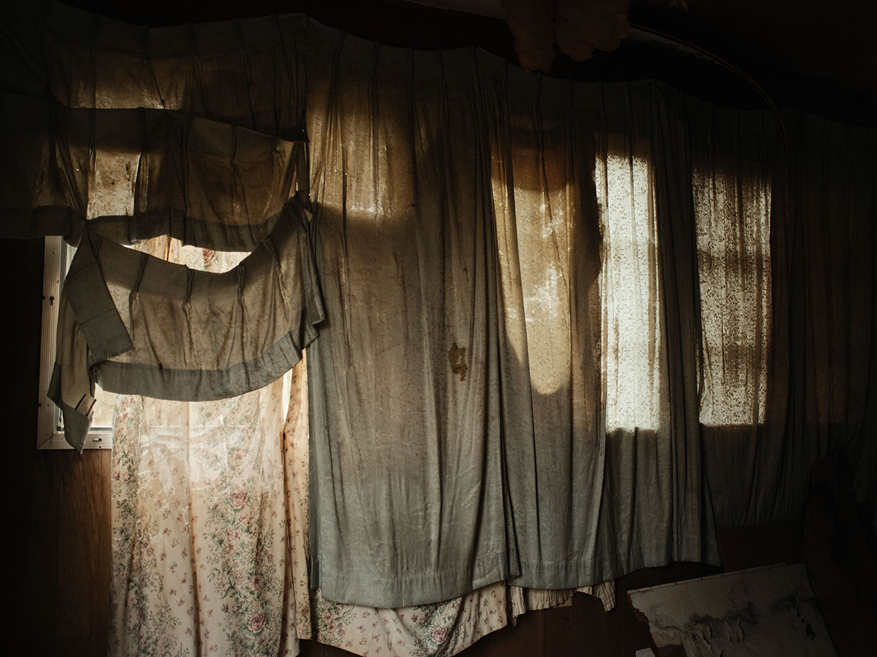 Sunlight illuminates layered floral and sheer curtains in a quiet, abandoned room at Lily of the Valley Campground,