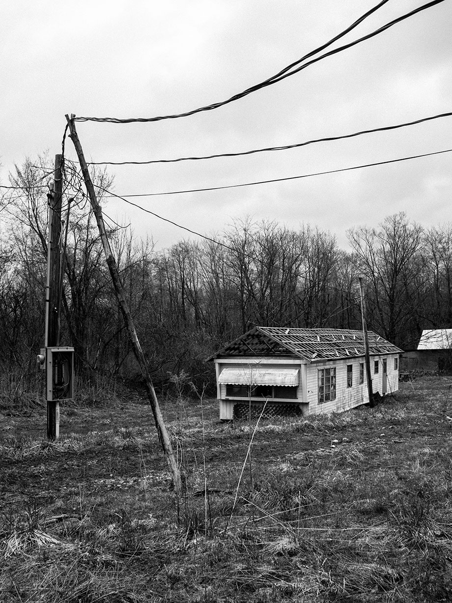 Black and white image of an abandoned, single-story building with a missing roof, alongside a utility pole and bare