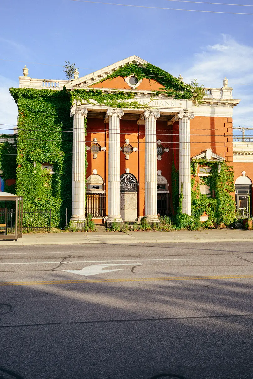 Abandoned Lake Shore Bank, a red brick building heavily overgrown with green ivy, featuring large white columns and a…